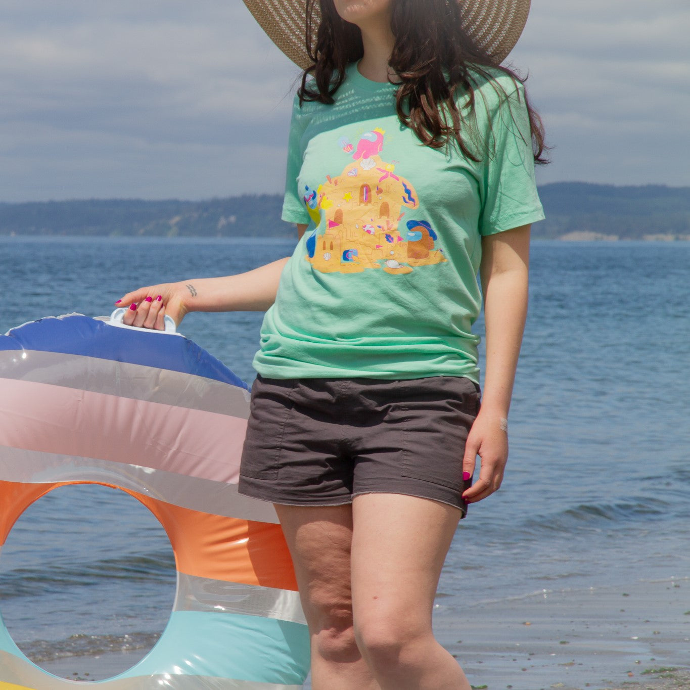  A product photo of the Among Us: Sandcastle Tee in Mint. The t-shirt is pistachio green and shows four crewmates building a sandcastle, which vaguely resembles the Airship. A pink crewmate wearing a crown hat sits at the top of the castle. 