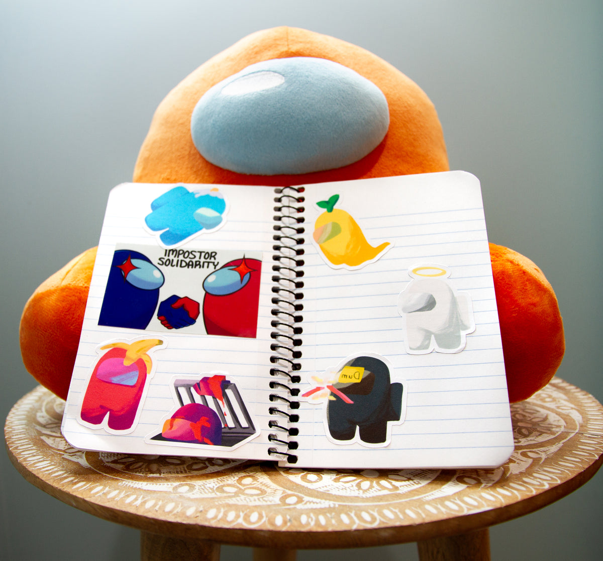 The notebook of Among Us stickers, six by Kuuji and one random, propped up against a sitting crewmate plush.
