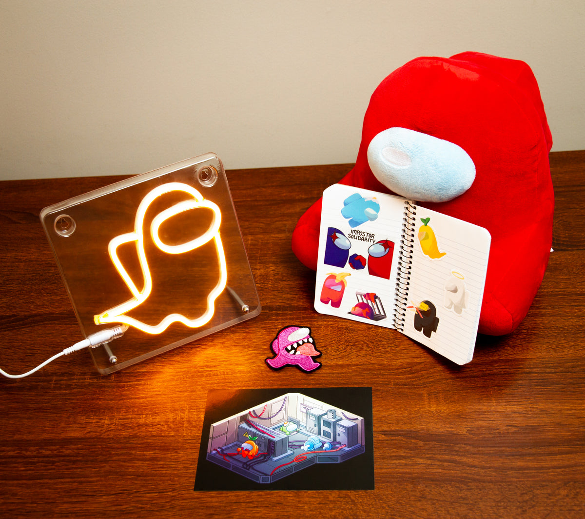 The entire Among Us Murder Room Artist Alley bundle of items arranged on a wooden desktop. The Ghost Crewmate LED is lit up on the left, a notebook covered in Kuuji stickers and one bonus sticker is on the right, and in the center lying flat is the purple Imposter patch and the Skeld Engineering print.