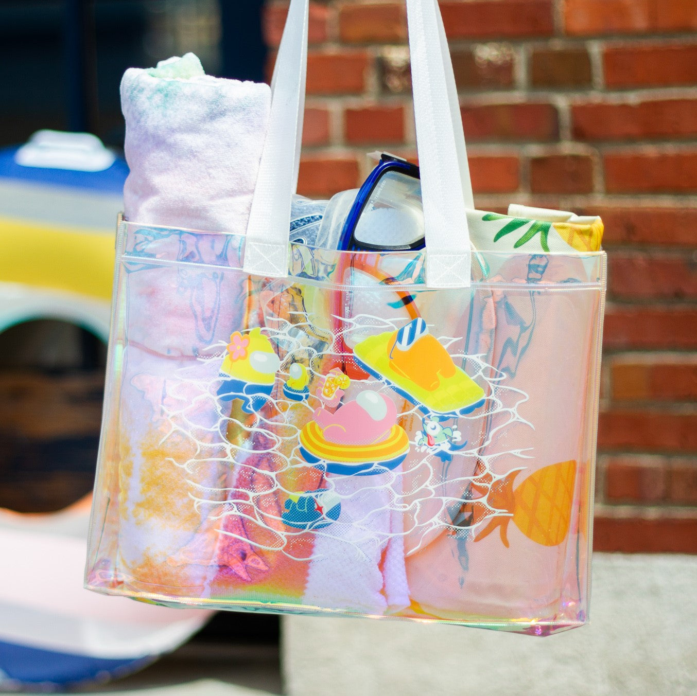 A product photo of the Among Us: Swimming Pool Tote. The tote is iridescent blue with natural canvas straps. The tote shows a pink crewmate lounging in an orange floatie. A large yellow crewmate observes a yellow mini crewmate, in a tiny floatie. An orange crewmate, wearing sunglasses, sun bathes on a big lime pool floatie.You can also see a snorkeling space dog and an impostor at the bottom of the pool. White lineart delineates ripples in the water.