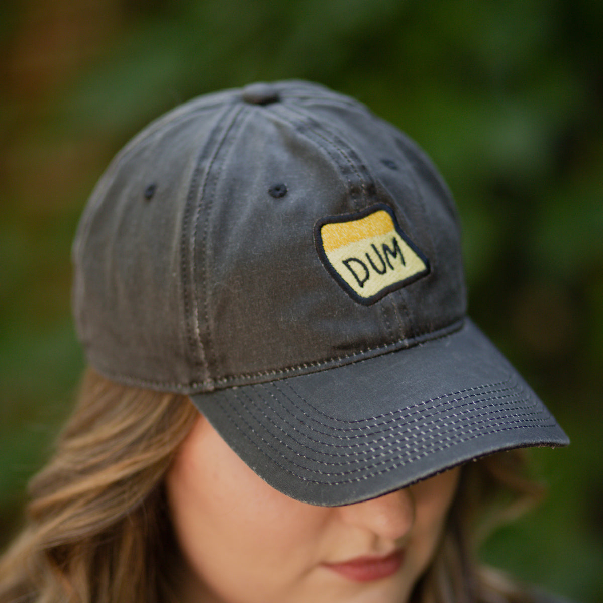 An overhead photograph of a model’s head looking down and to the right. She’s wearing a black DUM hat and you can see the embroidery detail on the yellow DUM sticky note.  