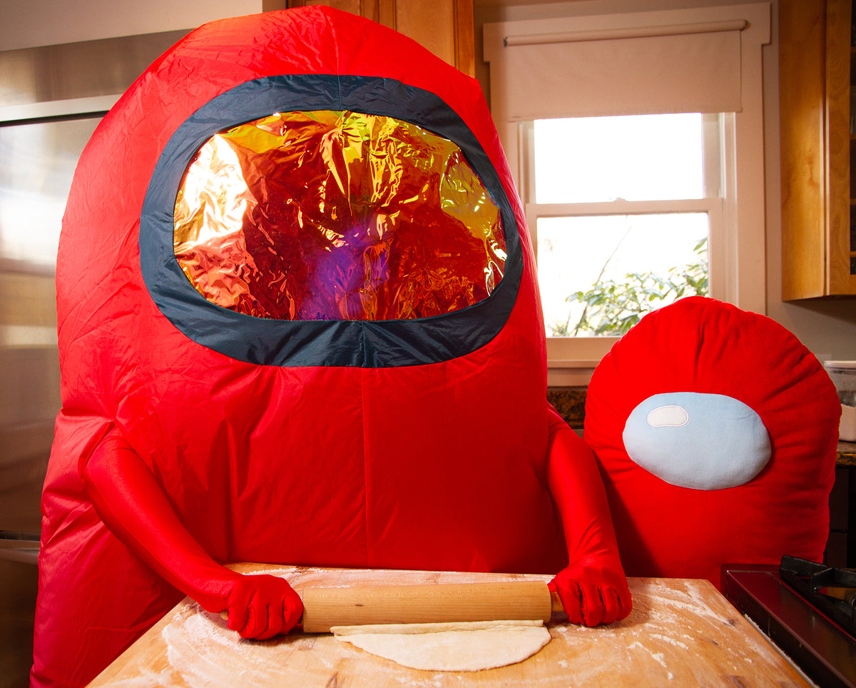 A Kitchen scene where a person is standing at the counter wearing the Red Crewmate Costume. They are using a light brown rolling pin to roll out some dough. Next to them is the Red Longbean Crewmate Plush standing next to them. 