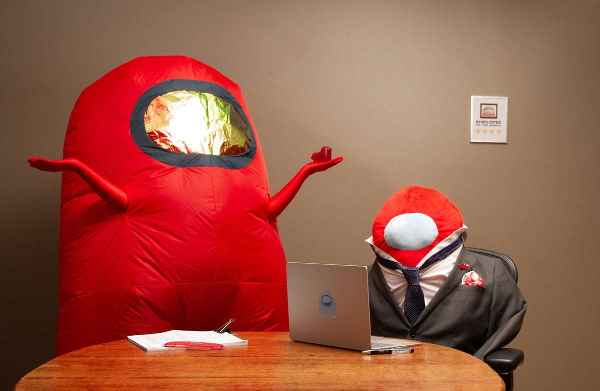 Red Crewmate Costume stands beside a table, holding up a Red Crewgi figure. In front of them is a stack of paperwork with a Crewmate ruler on top. Beside them is the Longbean Plush sitting at the table, looking at a laptop. Longbean wears a dark suit and navy tie. A blue Crewmate sticky note attached to the back of the laptop and the Among Us Floaty pen beside it. Behind Longbean is a placard that features another Red Crewmate and it says &quot;Employee of the Month&quot; with five yellow stars beneath it.