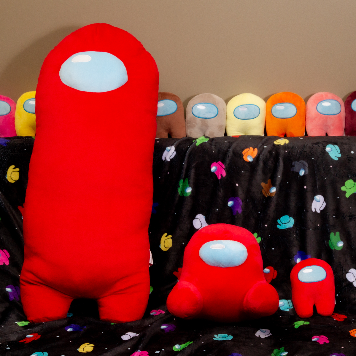 Several assorted colored tiny Crewmate Plush stand along the back of a couch. The Couch is draped with the patterned and colorful Crewmate Space Party Blanket. On top of the blanket is a line of three Red Crewmate Plush: a Longbean Plush, a Sitting Red Crewmate Plush, and a tinier Red Crewmate Plush. Longbean plush is designed by Zara Varin. The Sitting plush &amp; tinier plush are designed by Frisk Wolfie.