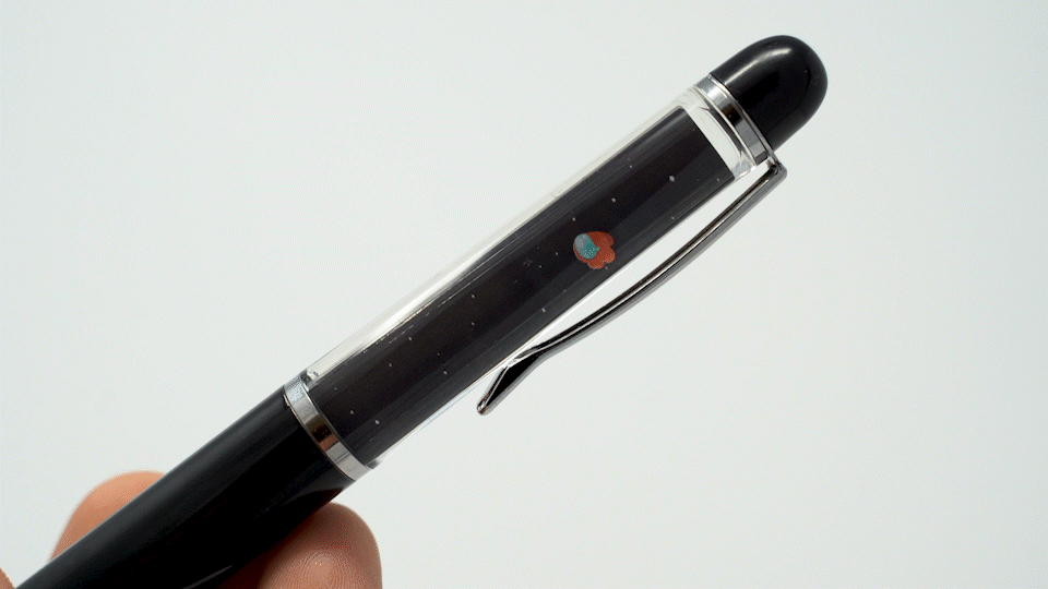 A black plastic floaty pen with a double-sided clear tube with Crewmate artwork by Noble Demons inside it. The back view of the clear tube features a red, crying, ejected Crewmate suspended in mineral oil liquid floating in space. A hand tilts the pen slowly showing the ejected Red Crewmate floating to the opposite side. 